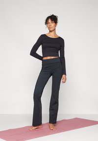 Crni dugih rukava top koji se završava iznad struka uparen s crnim širokim joggers hlačama. Model stoji bosi na ružičastom yoga prostirci. Glatka tkanina i uski kroj.