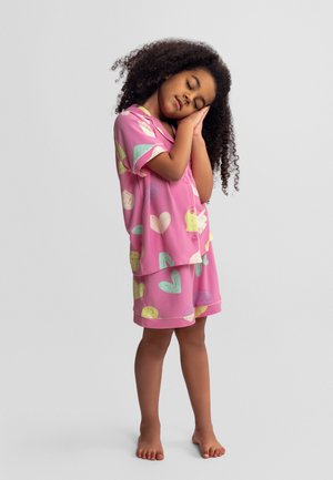 Kind met krullend haar in roze pyjama met hartjespatroon staat blootsvoets, ogen gesloten, handen tegen het gezicht gedrukt alsof het slaapt.