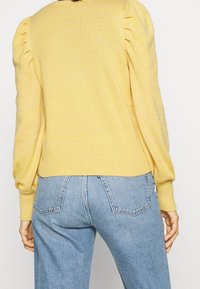 Pull en maille jaune à manches bouffantes et ourlet côtelé, associé à un jean taille haute bleu clair avec poches arrière et texture en denim.