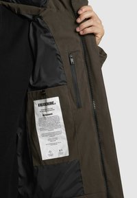 Smeđa vanjska jakna s crnom unutarnjom podstavom. Ima džep na rajsferšlus i oznaku za njegu. Jednostavan dizajn s teksturiranom završnom obradom.