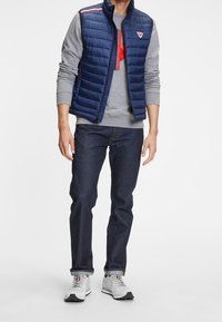 Marineblauer Puffer-Weste mit horizontalem Steppmuster über einem grauen Langarmshirt, kombiniert mit dunklen Jeans und grauen Sportschuhen.