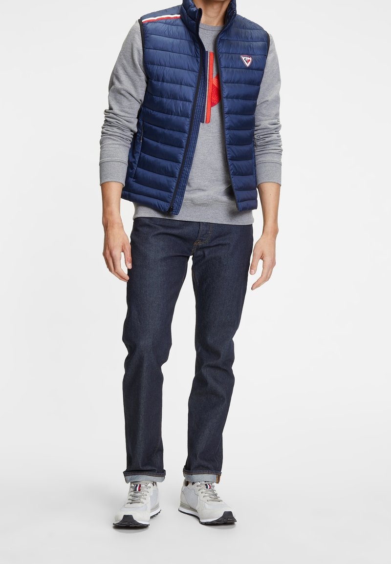 Marineblauer Puffer-Weste mit horizontalem Steppmuster über einem grauen Langarmshirt, kombiniert mit dunklen Jeans und grauen Sportschuhen.