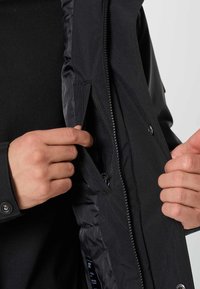 Mani che mostrano una tasca interna con zip sulla fodera interna nera di una giacca nera aperta.