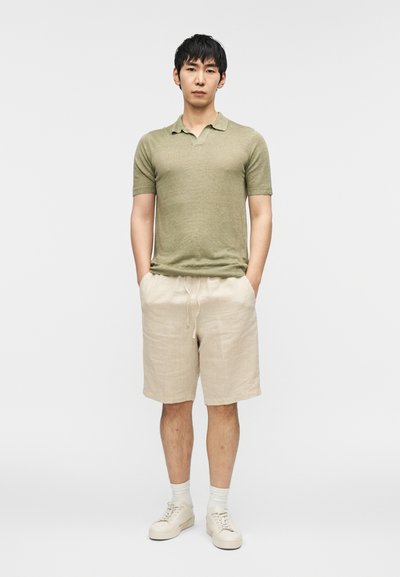 Polo vert, short en lin beige, chaussettes blanches, baskets légères. Le modèle se tient les mains dans les poches devant un fond uni.