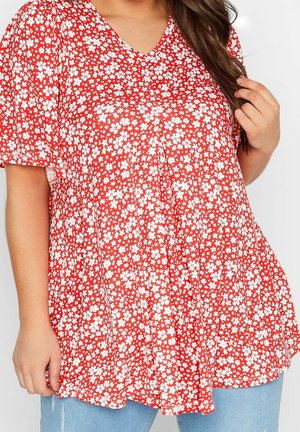 Kvinde iført rød kortærmet V-hals top med hvidt blomsterprint og lyseblå jeans, hånd rører ved håret og hviler ved siden.