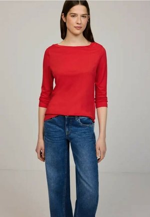 Giovane donna con indosso un top rosso a maniche lunghe e jeans blu, in piedi davanti a uno sfondo grigio chiaro uniforme.