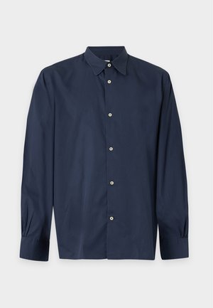 Homme portant des chaussures habillées noires, un pantalon brun foncé, une chemise bleu marine et un pull marron posé sur les épaules, debout, les mains dans les poches.