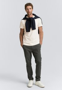 O ținută casual care include un tricou bej deschis și o pereche de pantaloni texturați gri închis, accesorizată cu un pulover bleumarin așezat pe umeri.