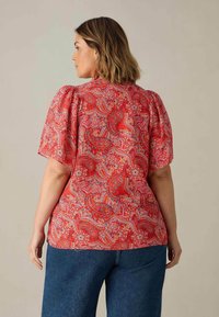 Rote Bluse mit Paisleymuster, kurzen Puffärmel und rundem Ausschnitt, aus leichtem Stoff, kombiniert mit blauen Jeanshosen.