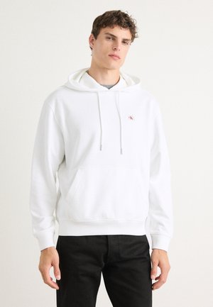 Jeune homme portant un sweat à capuche blanc avec un petit logo sur la poitrine et un pantalon noir, debout devant un fond clair uni.