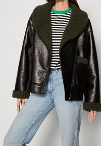 Veste en cuir noir avec col et poignets en polaire verte, fermeture éclair, poche avant, portée par-dessus une chemise à rayures et un jean clair.