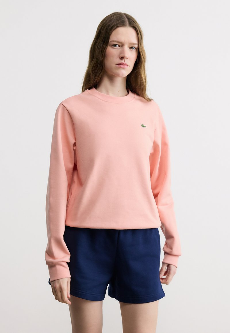 Νεαρή γυναίκα που φοράει ροζ μακρυμάνικη φούτερ και μπλε σκούρο σορτς, στέκεται μπροστά σε ένα απλό λευκό φόντο.