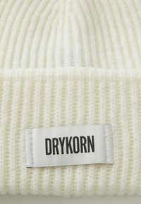Bijela rebrasta pletena kapa s oznakom od tkanine koja nosi naziv brenda "DRYKORN" u debelom crnom tekstu. Mekana tekstura, dizajn koji čvrsto prianja.