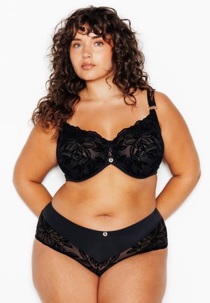 Donna con capelli ricci che indossa un reggiseno in pizzo floreale nero e slip in pizzo a vita alta abbinati, in piedi con le mani dietro la schiena.