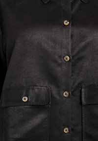 Primo piano di una camicia nera con bottoni, con quattro bottoni marroni e due tasche anteriori con patta, mostrando la texture del tessuto e i dettagli delle cuciture.