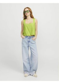 Zeleni laneni crop top s kvadratnim izrezom, svjetloplave široke traperice i bež papuče s dekorativnim detaljima. Model stoji s rukama u džepovima.