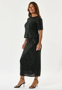 Must mustriga kaheosaline komplekt, millel on lühikesed varrukad ja põlve pikkusega seelik, koos mustade rihmadega kontsadega. Sile, läikiv tekstuur.