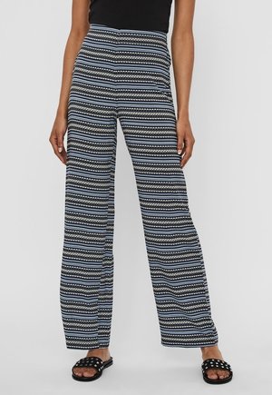 Vrouw draagt een wijde broek met hoge taille in zwart-wit-blauw patroon, zwarte studs slippers en een zwarte top.