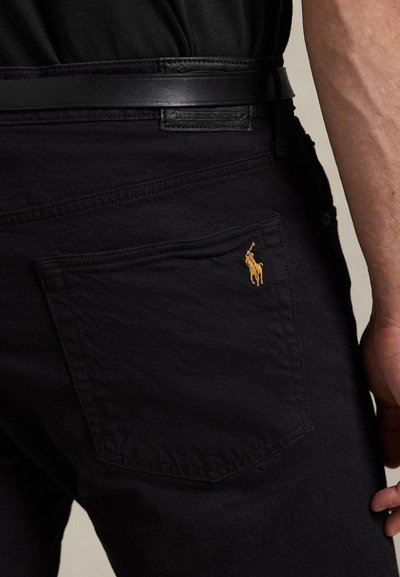 Close-up of a person wearing black jeans with a brown embroidered horse logo on the back pocket and a black leather belt.