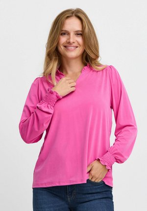 Blusa a maniche lunghe rosa con scollatura arricciata e polsini smockati. Tessuto morbido e liscio con vestibilità ampia e spacco laterale per una maggiore libertà di movimento.