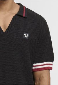Polo negro de punto con escote en V, ribete rojo en el collar y mangas cortas a rayas con acentos rojos y blancos. Incluye un pequeño parche con el logotipo.