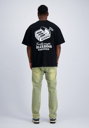 Mann von hinten, trägt ein schwarzes T-Shirt mit einer Sardinendosengrafik und dem Text "Freshly caught BLCKBNNS SARDINES", helle Jeans, schwarze Schuhe.