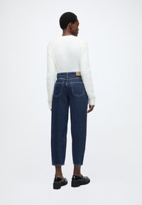 Άσπρο αφράτο πουλόβερ, ψηλόμεσο σκούρο μπλε τζιν και μαύρα παπούτσια πλατφόρμα. Το τζιν έχει κλασικό σχέδιο τσέπης και ανοιχτό χρώμα ραφών.