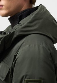 Padded jacket w ciemnooliwkowym kolorze, z kapturem, teksturowanym materiałem oraz kieszenią klapkową z naszywką na rękawie.