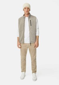 Graue Fleece-Weste mit einem Reißverschluss vorne, darunter ein weißes Langarmshirt, beigefarbene Hose und eine cremefarbene Strickmütze. Weiße Turnschuhe runden das Outfit ab.