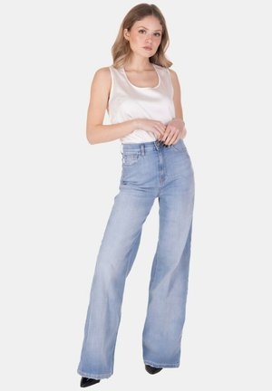 Giovane donna che indossa un top bianco senza maniche e jeans a vita alta azzurri chiari a gamba larga, in piedi con le mani vicino alla vita su sfondo bianco.