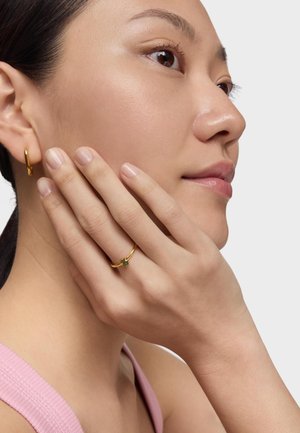Gold hoop earrings and a gold ring with a green gemstone, worn by a person with a smooth complexion and long hair. Light pink top visible.