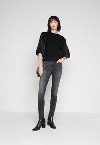 Fekete, bordás felső, redőzött bőrujjakkal, szürke skinny farmerral és fekete sarkú bokacsizmával. Egyszínű szürke háttér előtt.
