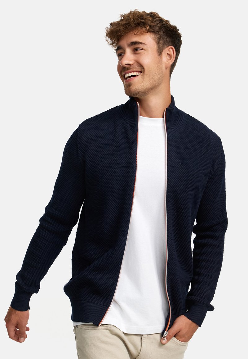 Cárdigan azul marino con cremallera, fabricado en punto texturizado, cuello alto, ribetes rojos en contraste y puños acanalados; llevado sobre una camiseta blanca.