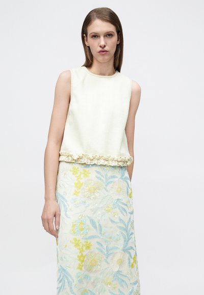 Haut blanc sans manches avec ourlet à volants, associé à une longue jupe ornée d'une broderie florale bleue et jaune sur un fond clair.