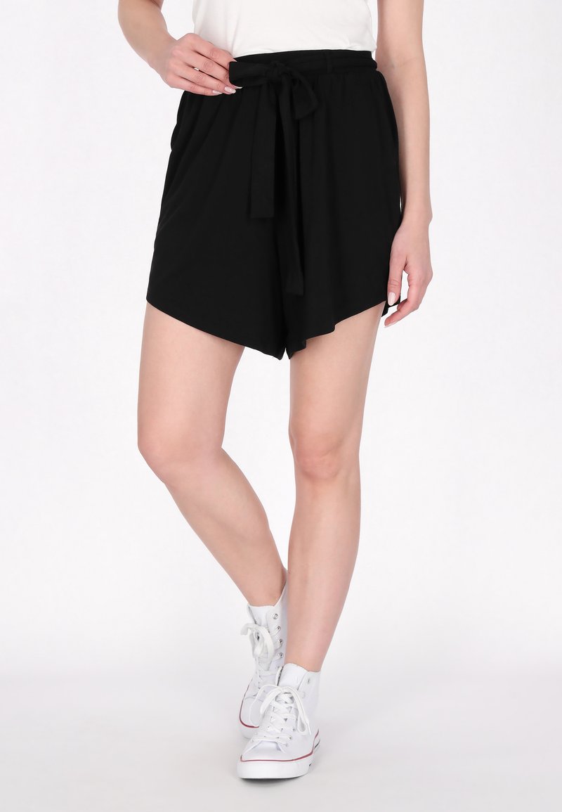 Schwarze Shorts mit einem gebundenen Gürtel, locker geschnitten und mit abgerundetem Saum. Das Material wirkt weich mit einer glatten Textur. Dazu werden weiße Sneakers getragen.