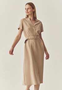 Satin-beiges Kleid mit kurzen Ärmeln, V-Ausschnitt, tailliertem Bund und knielangem Saum. Verfügt über eine Seitentasche und eine glatte, elegante Textur.