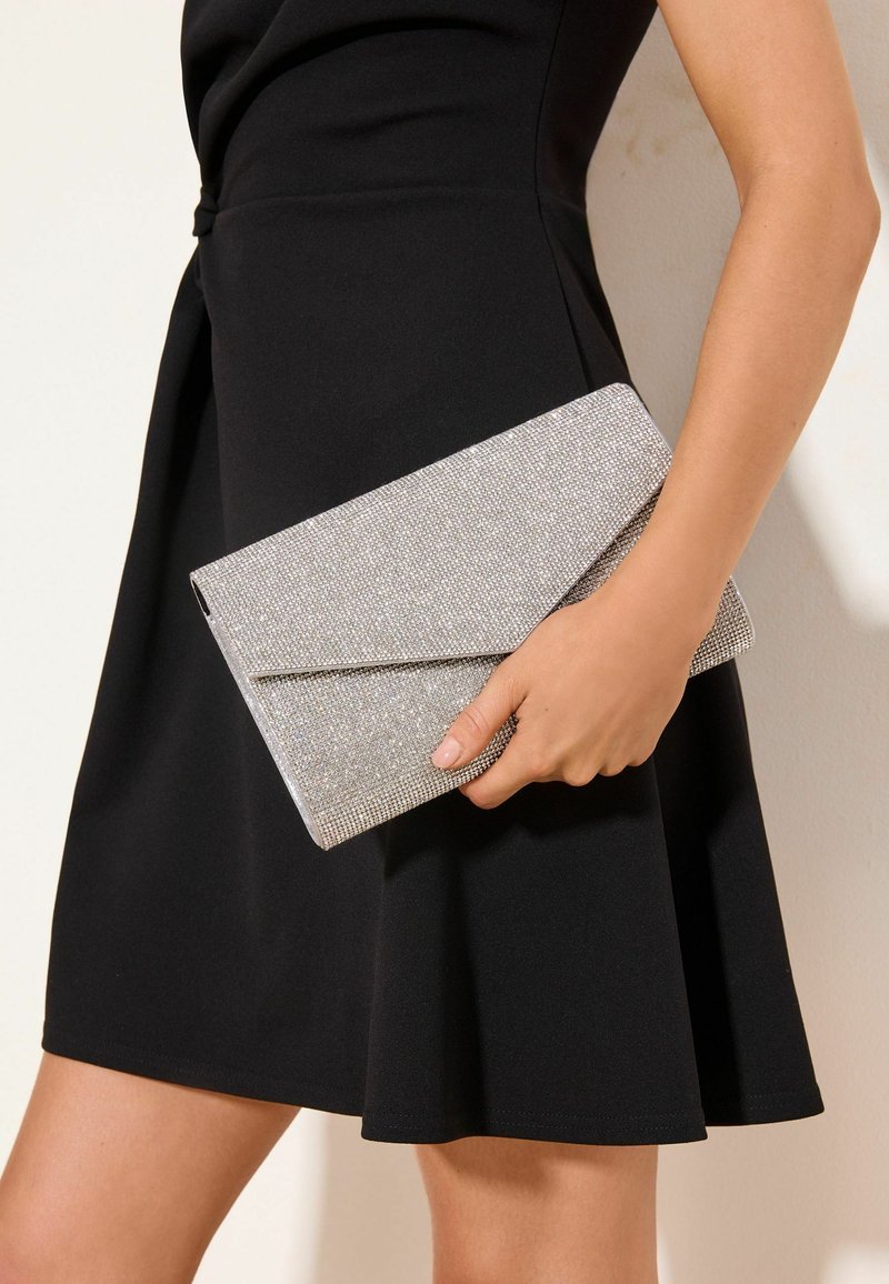 Moteris juodoje suknelėje laiko sidabrinę, žėrinčią aplanko formos rankinę šviesiame fone.