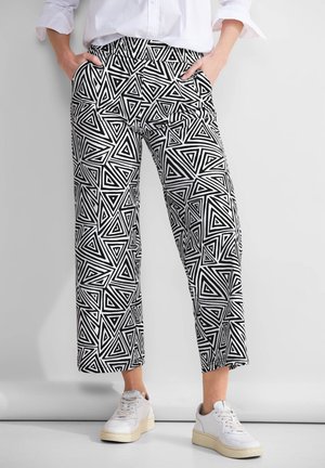 Personne portant un pantalon à motifs géométriques noir et blanc, des baskets blanches, et une chemise blanche à manches longues, les mains dans les poches.