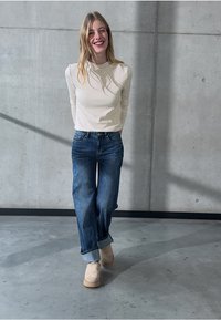 Lichtbeige langemouwen top, blauwe wijde jeans met opgerolde manchetten en beige instappers. Gladde textuur en casual pasvorm. Betonnen achtergrond.