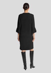 Schwarzes Kleid mit Glockenärmeln, knielang, aus glattem Stoff. Kombiniert mit hohen schwarzen Lederstiefeln. Schlichtes, minimalistisches Design.