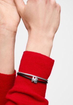 Muñeca izquierda con pulsera de dijes de mariposa en negro y plata, usada sobre un suéter rojo de manga larga.