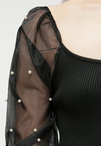 Um close-up de uma blusa preta com texturas em relevo, com uma mangas bufantes transparentes adornadas com pequenas pérolas brancas, sobre um fundo de ombro nu.