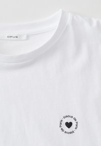 Wit katoenen T-shirt met een ronde hals en korte mouwen. Voorzien van een zwarte cirkelvormige print met tekst en een hartontwerp op de borst.