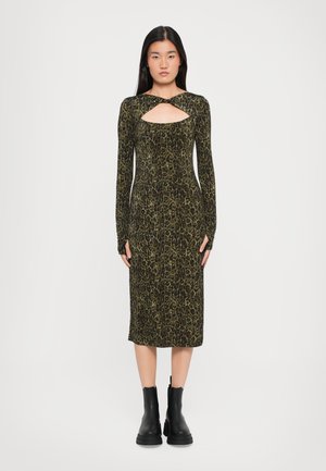 Mouwloze jurk met een luipaardprint in donkergroen en zwart, met een uitsnijding aan de voorkant. Draagt zwarte enkellaarsjes.