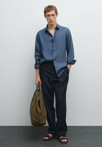Blue, long-sleeve button-up shirt with rolled cuffs, paired with dark pinstriped pants. A green canvas bag is held in one hand. Brown sandals worn.