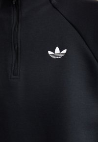 Sweatshirt noir avec un col montant et une demi-fermeture éclair, arborant un logo brodé blanc avec trois feuilles et des bandes horizontales.