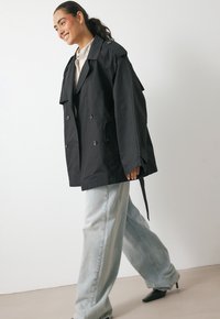 Young woman smiling, walking, wearing oversized black trench coat, light blue wide-leg jeans, beige shirt, and black pointed-toe heels.
