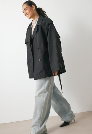 Young woman smiling, walking, wearing oversized black trench coat, light blue wide-leg jeans, beige shirt, and black pointed-toe heels.