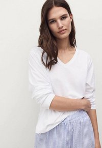Camicia bianca a maniche lunghe con scollatura a V, realizzata in tessuto morbido, abbinata a pantaloni a righe blu chiaro. La camicia ha le maniche arrotolate e una vestibilità rilassata.