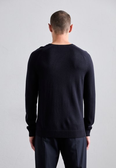 Homme aux cheveux courts portant un pull noir à manches longues et un pantalon sombre, se tenant de dos face à l'appareil photo devant un mur gris uni.